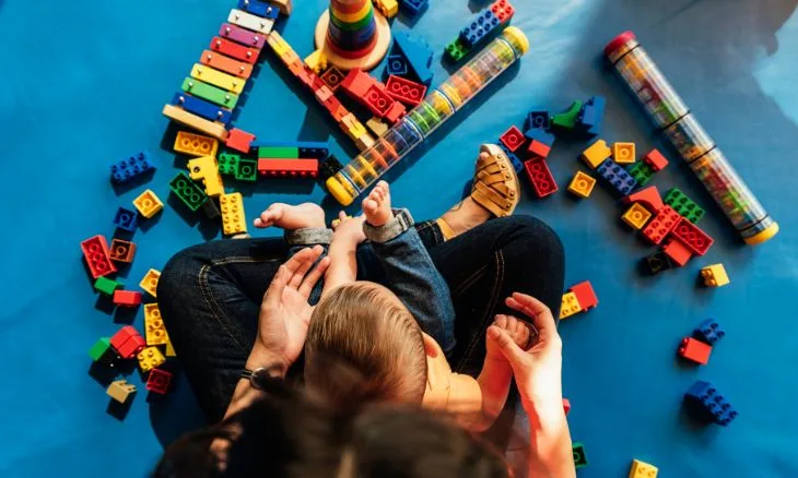 Brinquedos espalhados pelo chão, mãe com criança no colo
